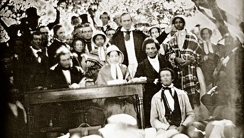 The 1850 Fugitive Slave Law Convention, Cazenovia, New York. The Edmonson sisters are standing wearing bonnets and shawls in the row behind the seated speakers. Frederick Douglass is seated, with Gerritt Smith standing behind him, and with Abby Kelley Foster the likely person seated on Douglass's left.